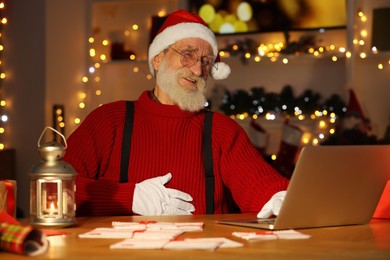 Santa Claus using laptop and laughing at his workplace in room decorated for Christmas