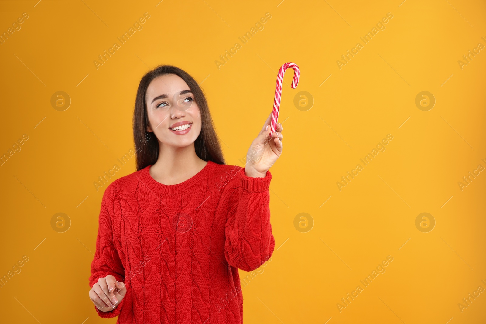 Photo of Young woman in red sweater holding candy cane on yellow background, space for text. Celebrating Christmas