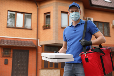 Photo of Courier in protective mask and gloves with pizza boxes near house outdoors. Food delivery service during coronavirus quarantine