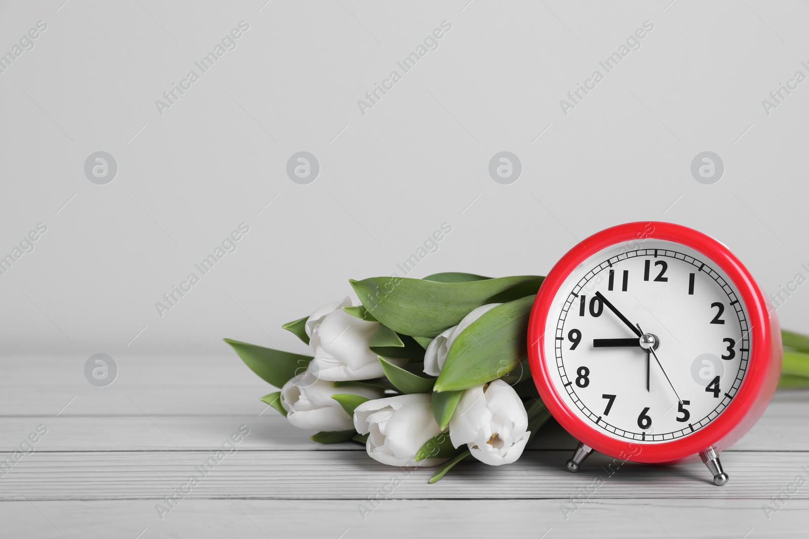 Photo of Red alarm clock and beautiful tulips on white wooden table against light background, space for text. Spring time