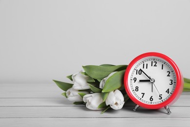 Photo of Red alarm clock and beautiful tulips on white wooden table against light background, space for text. Spring time