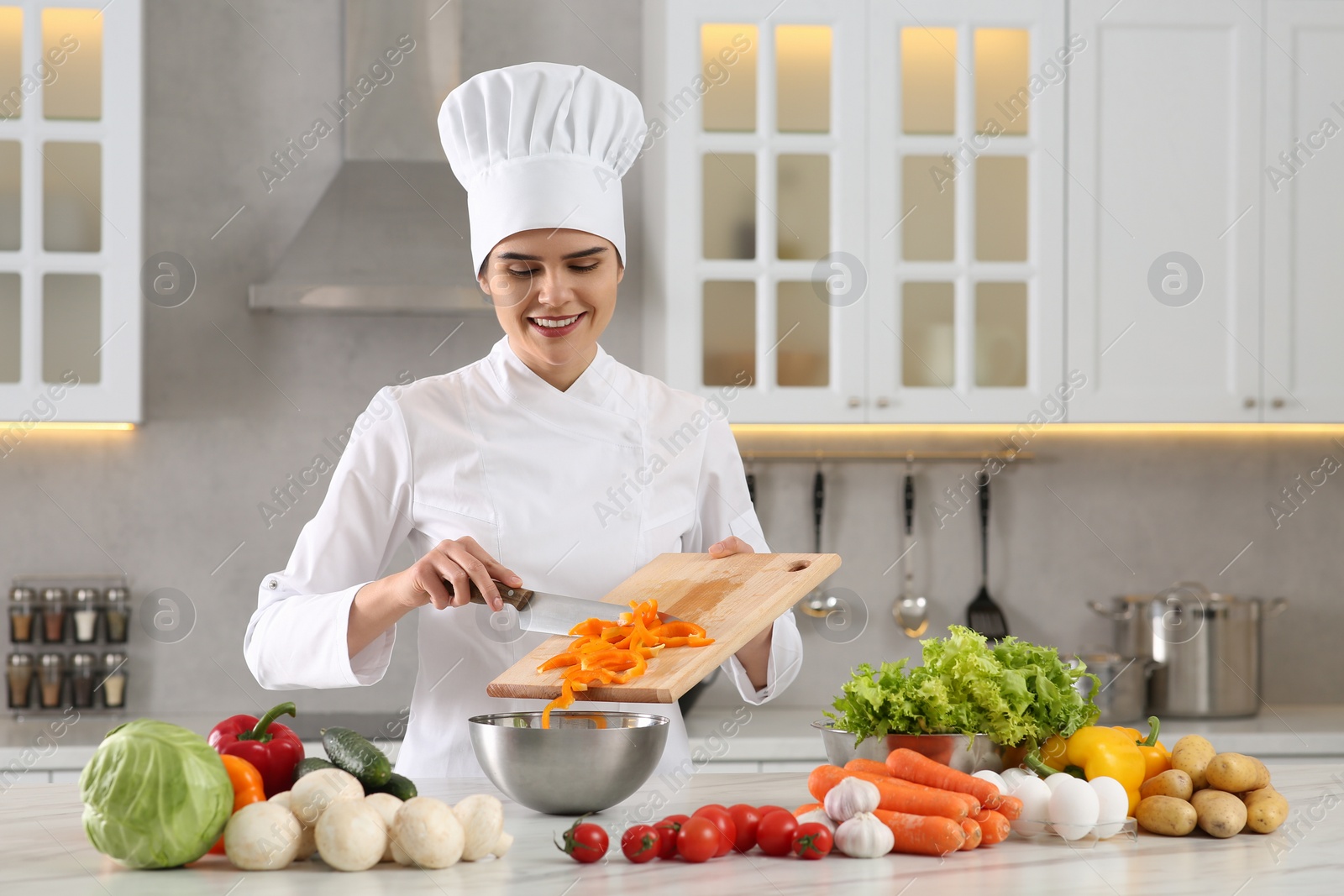 Photo of Professional chef putting cut bell pepper into metal bowl at white marble table in kitchen