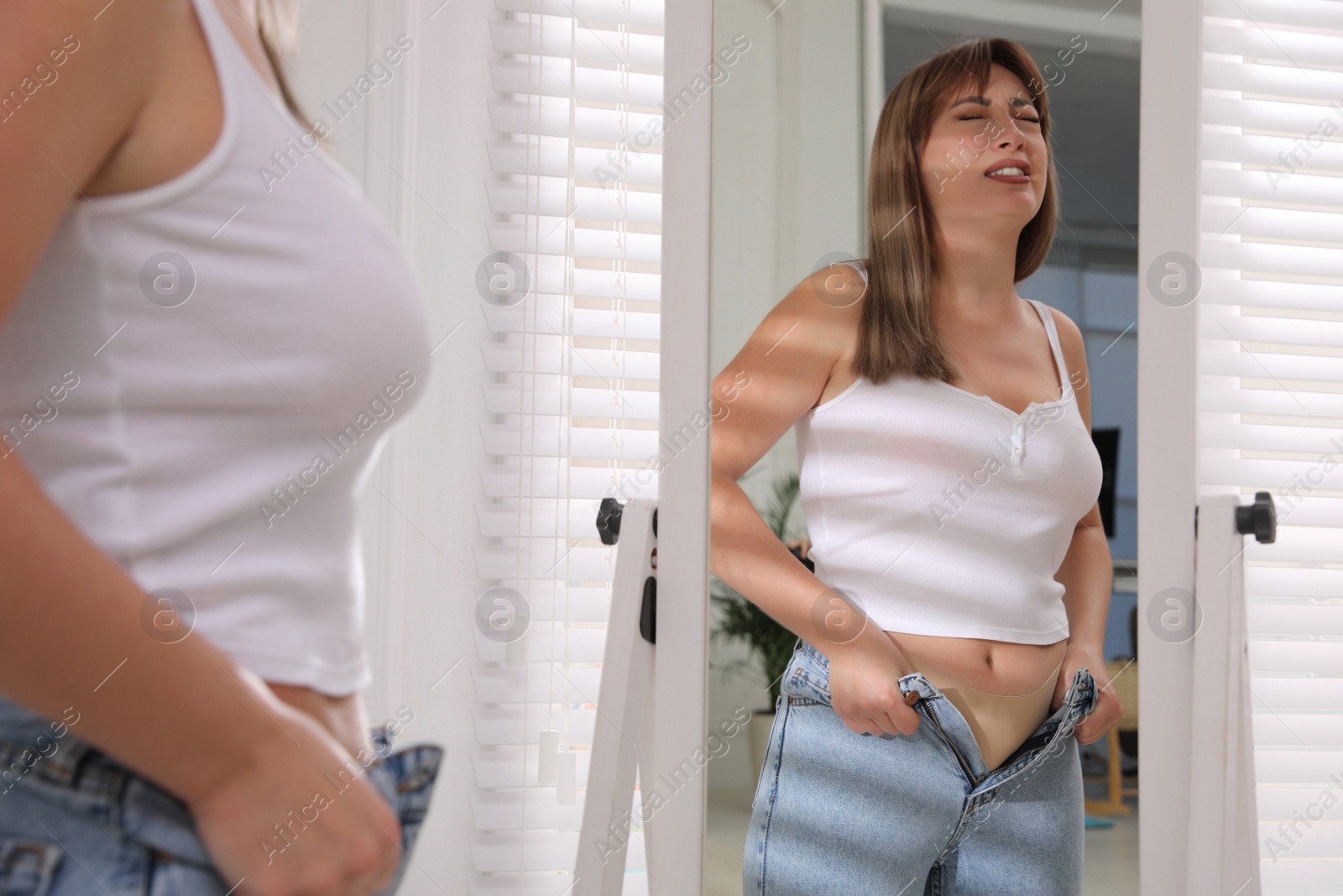 Photo of Young woman struggling to put on tight jeans near mirror at home
