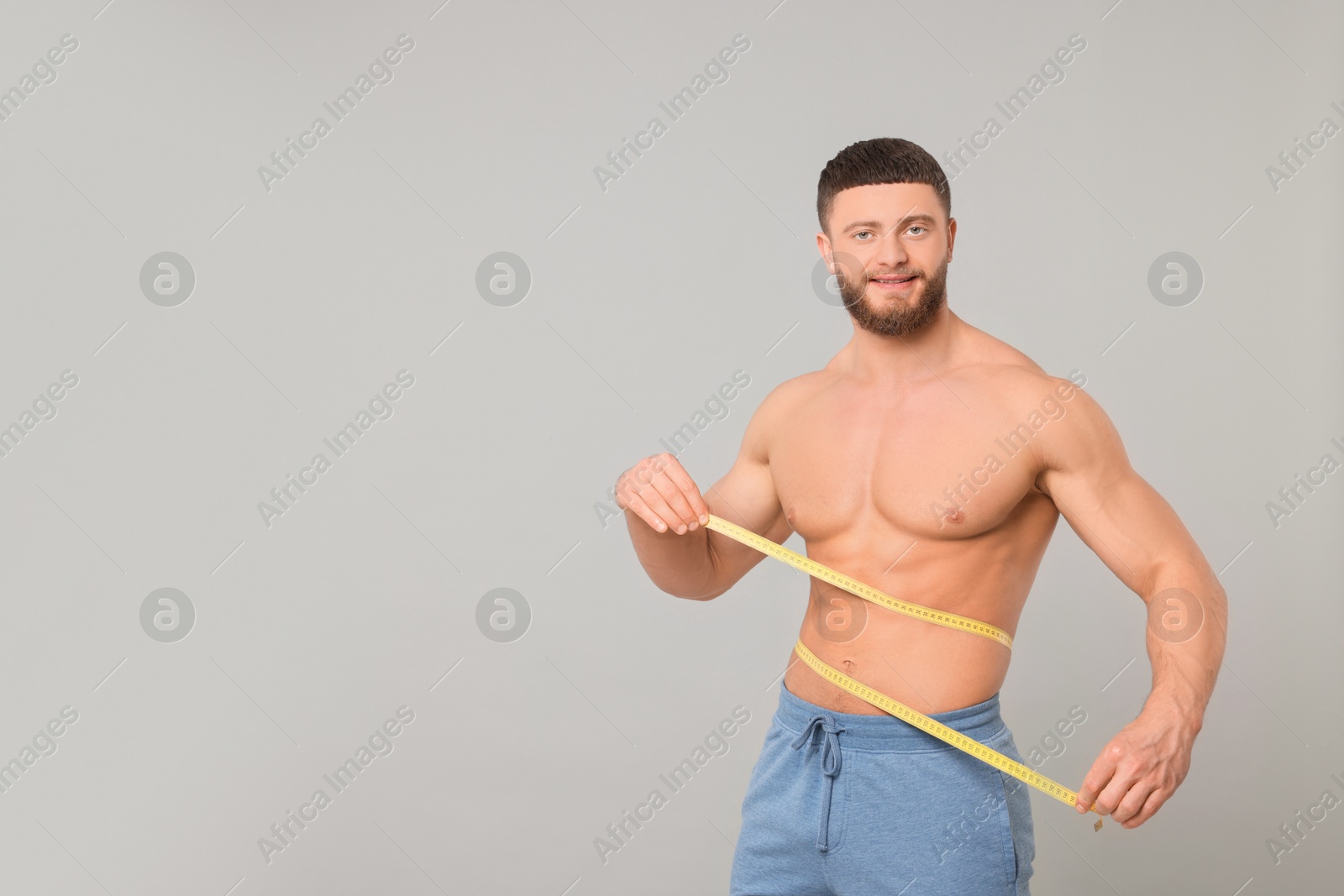 Photo of Portrait of happy athletic man measuring waist with tape on light grey background, space for text. Weight loss concept