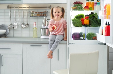 Cute girl with apple sitting near refrigerator in kitchen