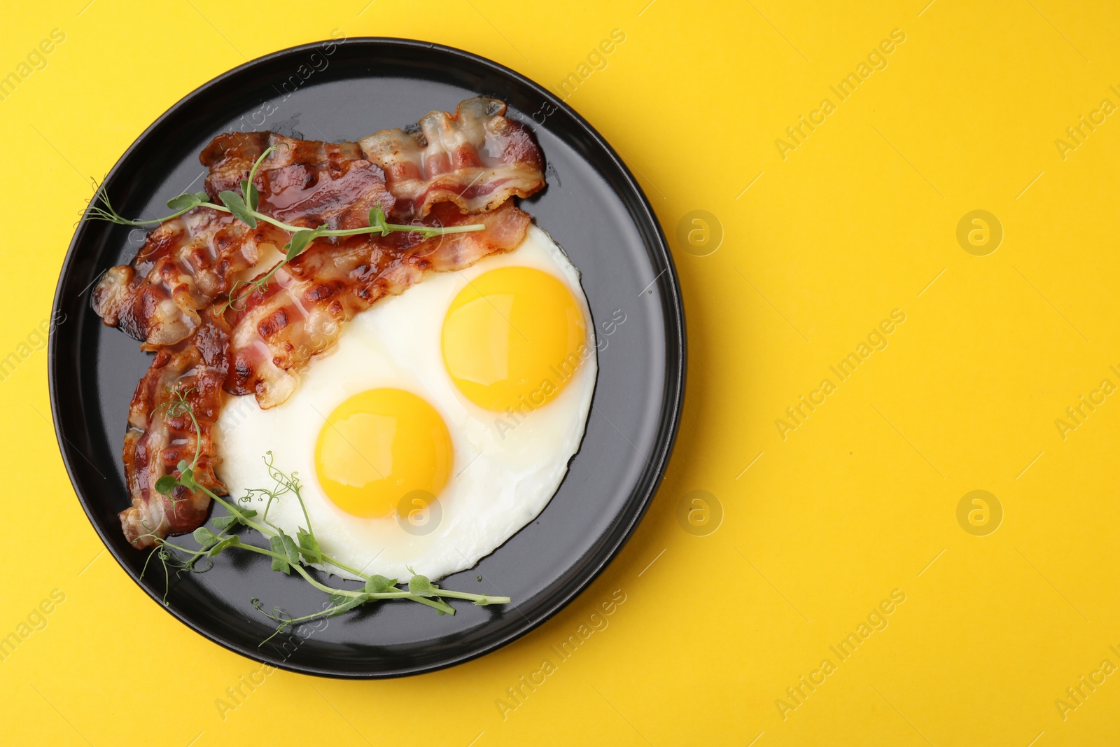 Photo of Fried eggs, bacon and microgreens on yellow background, top view. Space for text