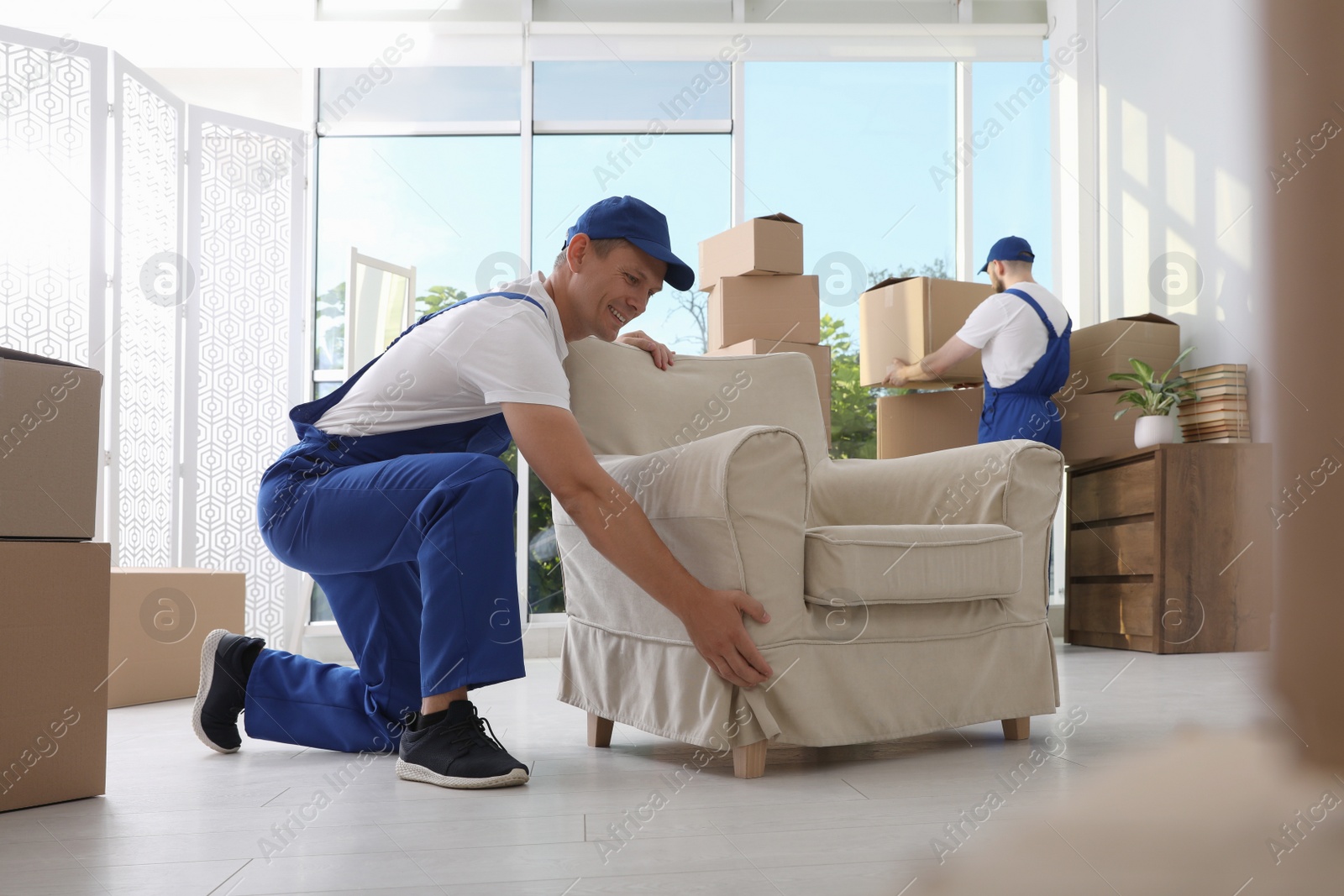 Photo of Moving service employees with armchair and cardboard boxes in room