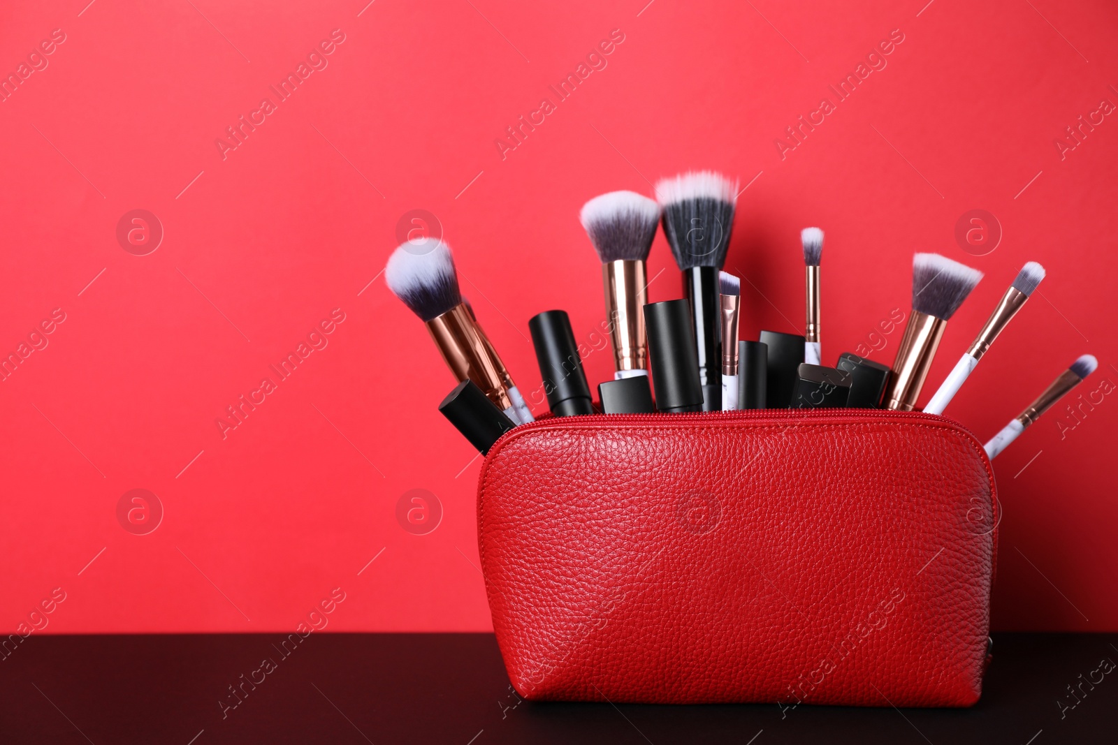 Photo of Bag with makeup brushes and cosmetic products on black table against red background. Space for text