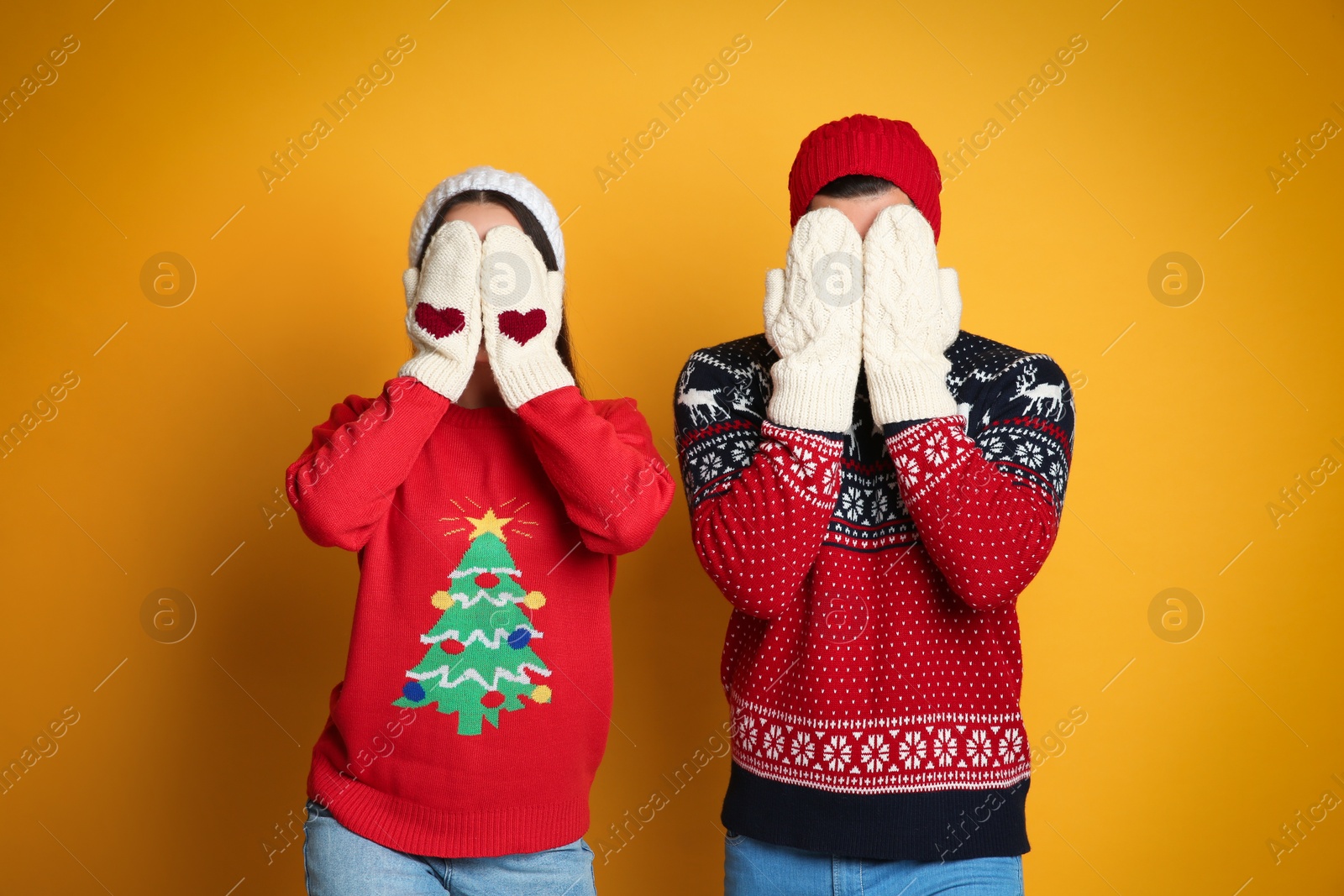 Photo of Couple in Christmas sweaters, knitted mittens and hats hiding faces on yellow background