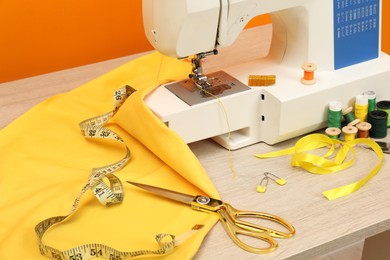 Photo of Modern sewing machine with cloth and craft accessories on wooden table near orange wall, closeup