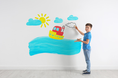 Little boy drawing ship and sea on white wall indoors