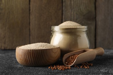 Jar and bowl of buckwheat flour near scoop with grains on black table against wooden wall