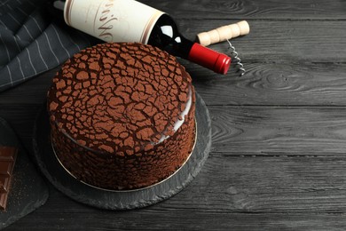 Photo of Delicious truffle cake, bottle of wine, chocolate pieces and corkscrew on black wooden table, space for text