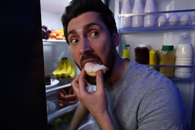 Photo of Man eating donut near refrigerator in kitchen at night. Bad habit