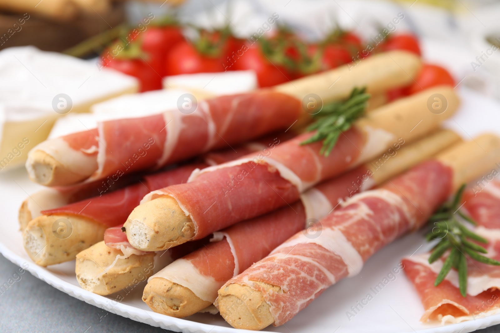 Photo of Plate of delicious grissini sticks with prosciutto, cheese and tomatoes on light grey table, closeup