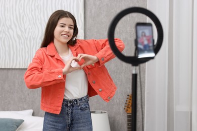 Smiling teenage blogger making heart gesture to her subscribers while streaming at home