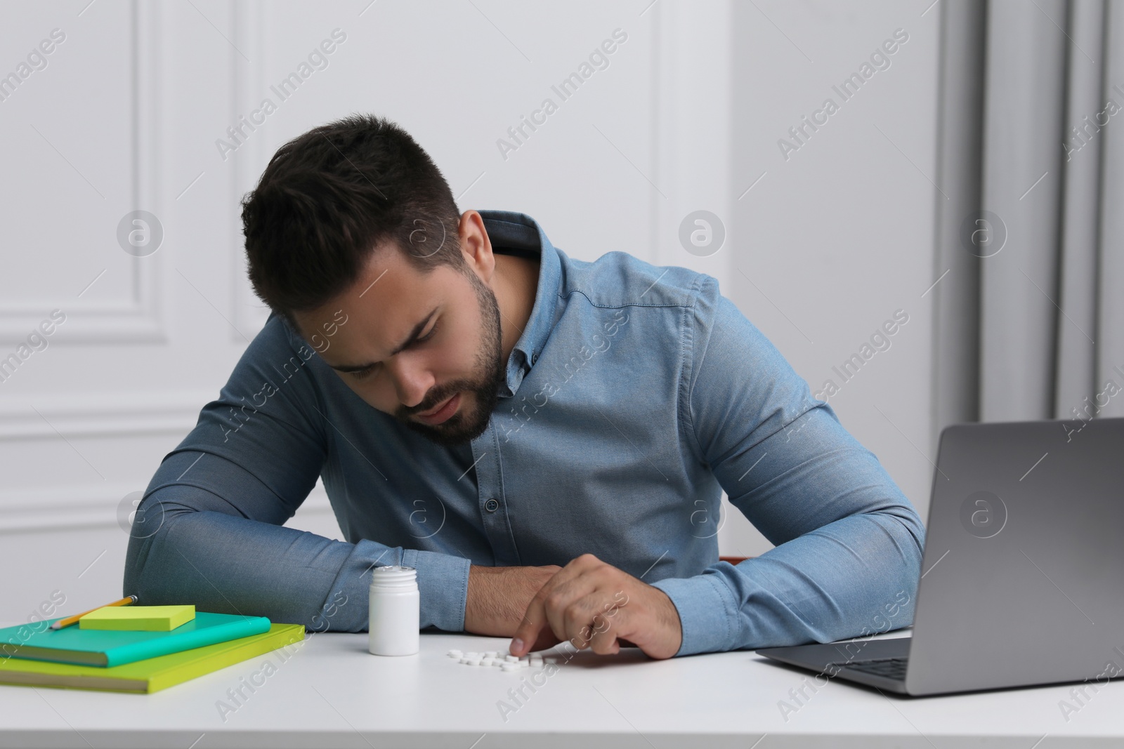 Photo of Depressed man at white table with antidepressants, laptop and stationery indoors