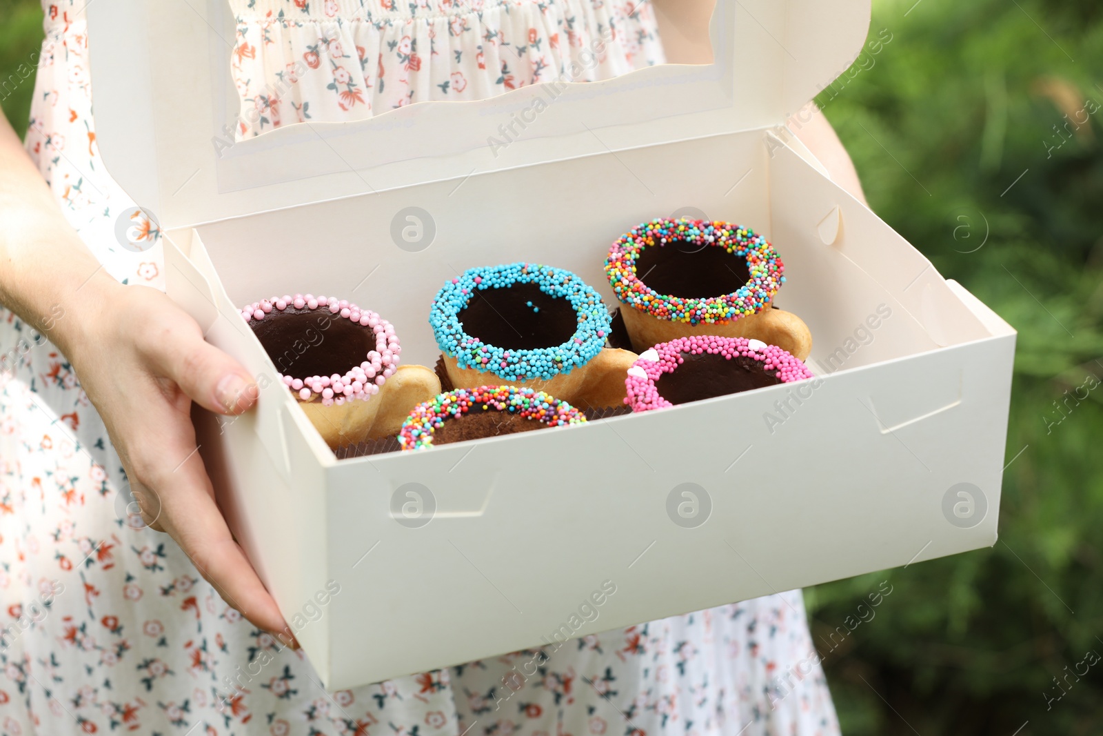 Photo of Woman holding box of delicious edible biscuit coffee cups decorated with sprinkles outdoors, closeup