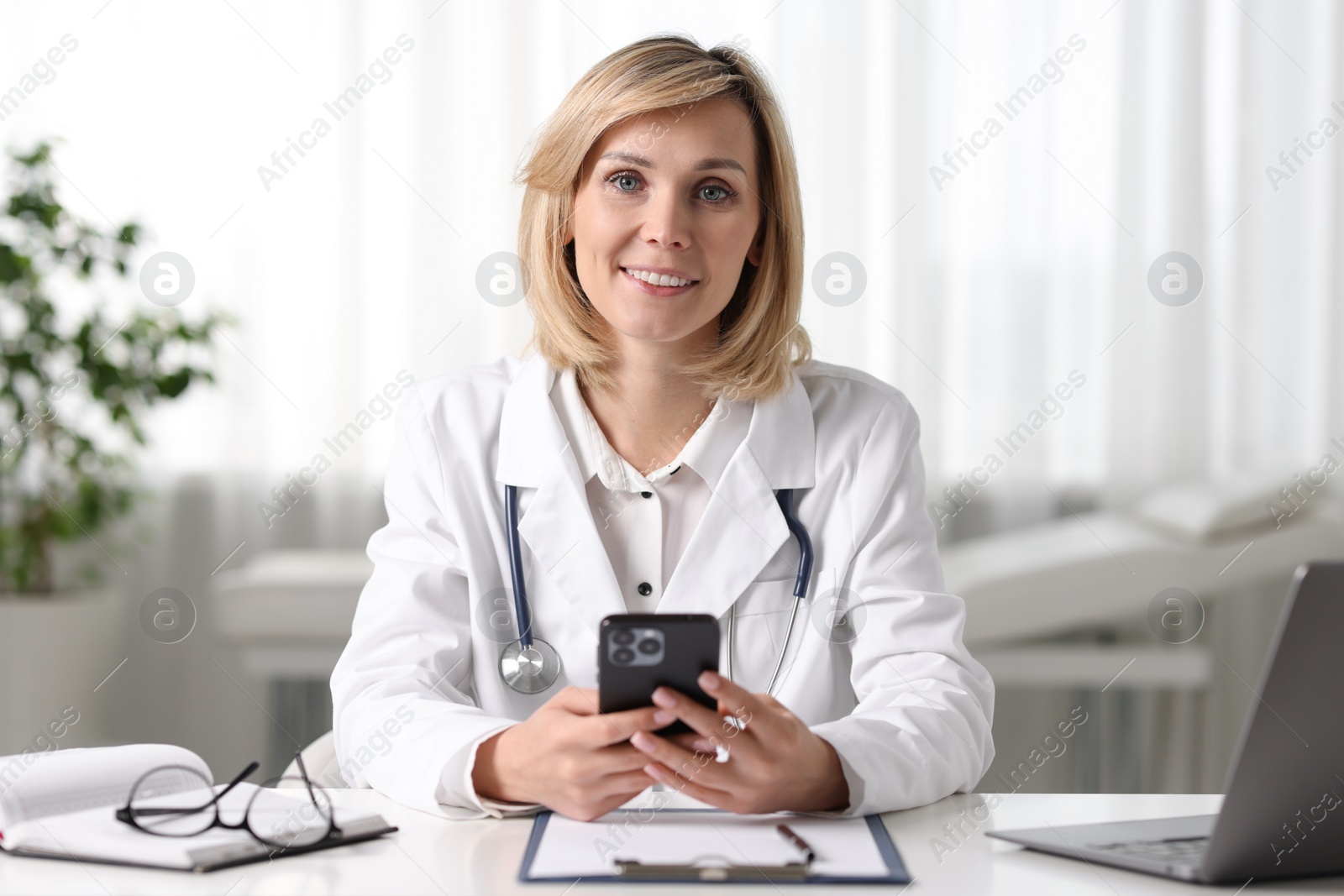 Photo of Smiling doctor with smartphone having online consultation at table in office