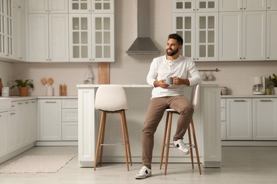 Photo of Handsome young man with cup of tea sitting on stool in kitchen