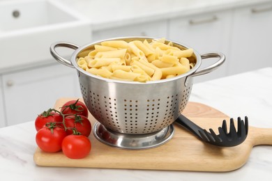 Photo of Cooked pasta in metal colander and tomatoes on light marble table, closeup
