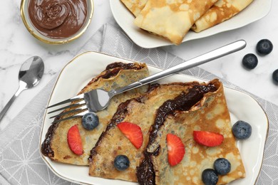 Photo of Tasty crepes with chocolate paste and berries served on white marble table, flat lay