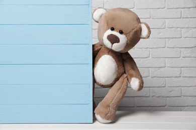 Cute teddy bear peeking out behind light blue wooden wall against brick background, space for text