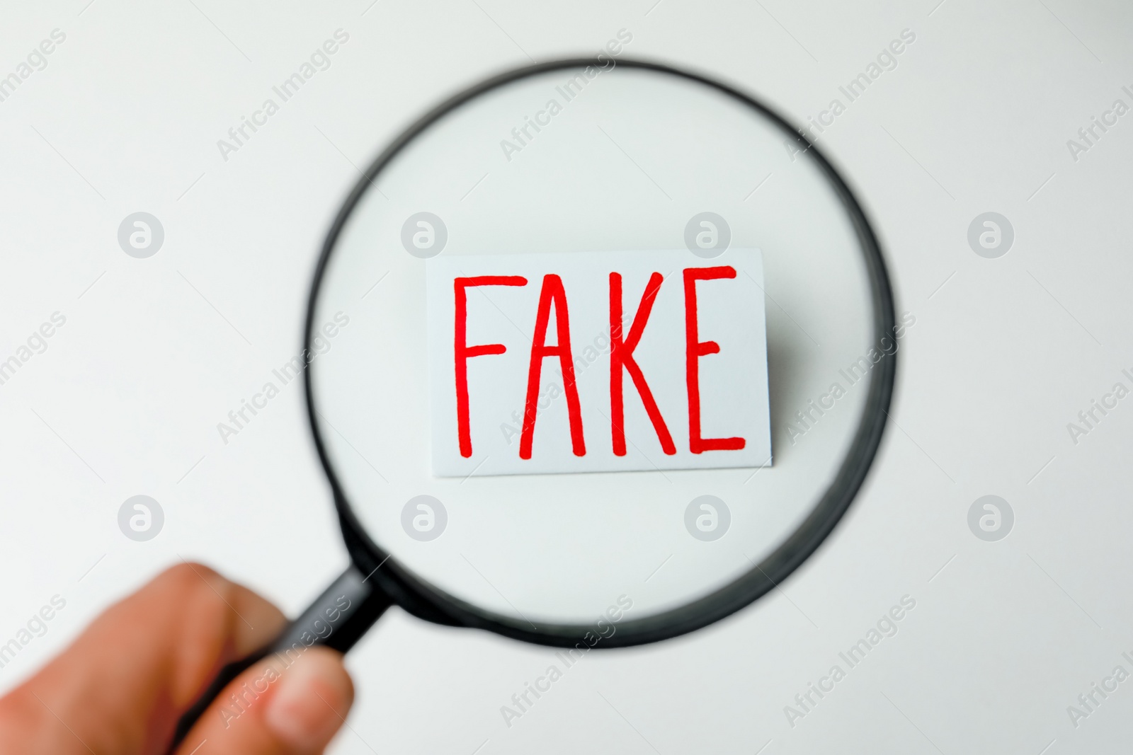 Photo of Woman holding magnifying glass over sheet of paper with word Fake on light background, top view