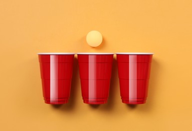 Plastic cups and ball for beer pong on orange background, flat lay