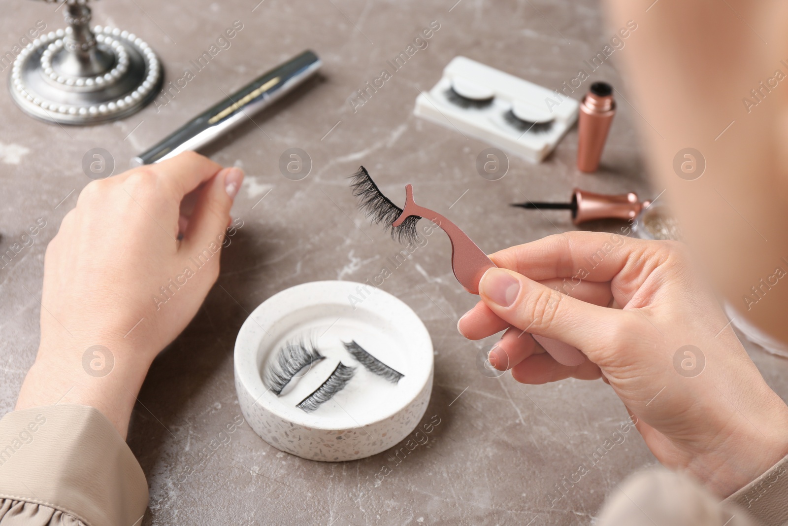 Photo of Woman holding tweezers with magnetic eyelashes at grey table, closeup
