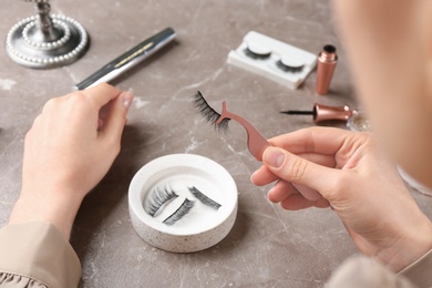 Photo of Woman holding tweezers with magnetic eyelashes at grey table, closeup