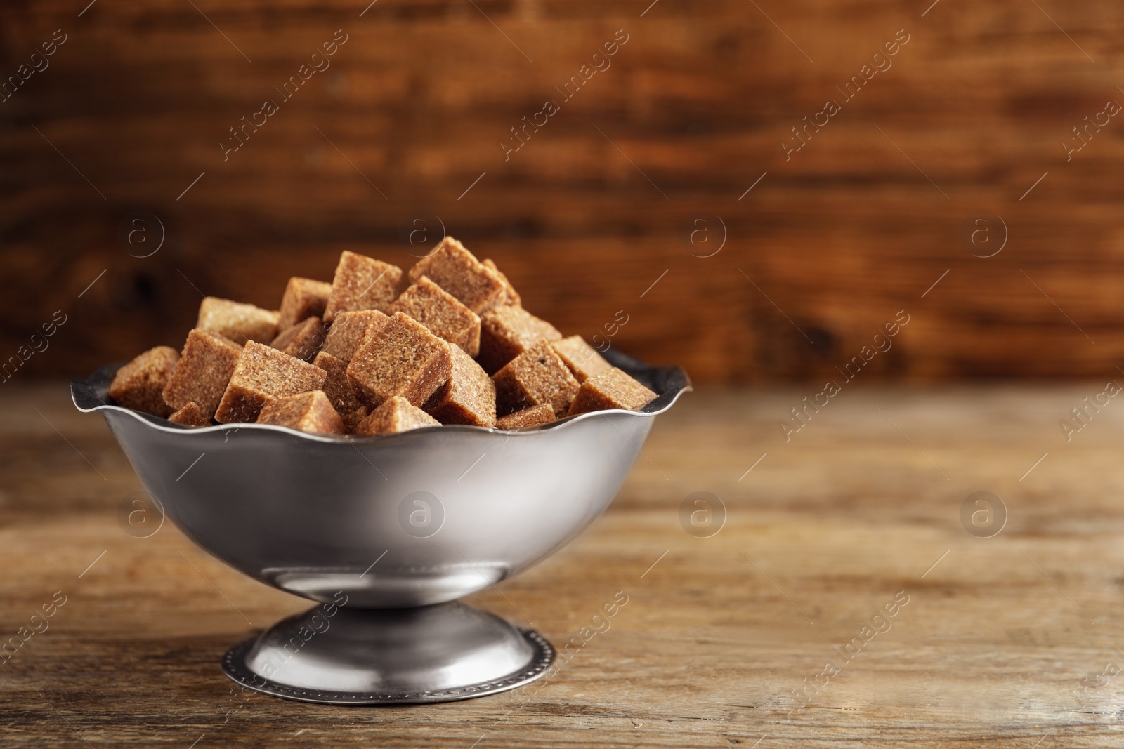 Photo of Brown sugar cubes in metal bowl on wooden table. Space for text