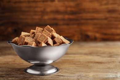 Photo of Brown sugar cubes in metal bowl on wooden table. Space for text