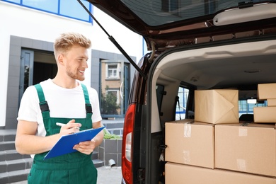 Photo of Young courier holding clipboard near delivery van with parcels outdoors