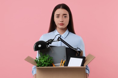 Photo of Unemployment problem. Confused woman with box of personal office belongings on pink background