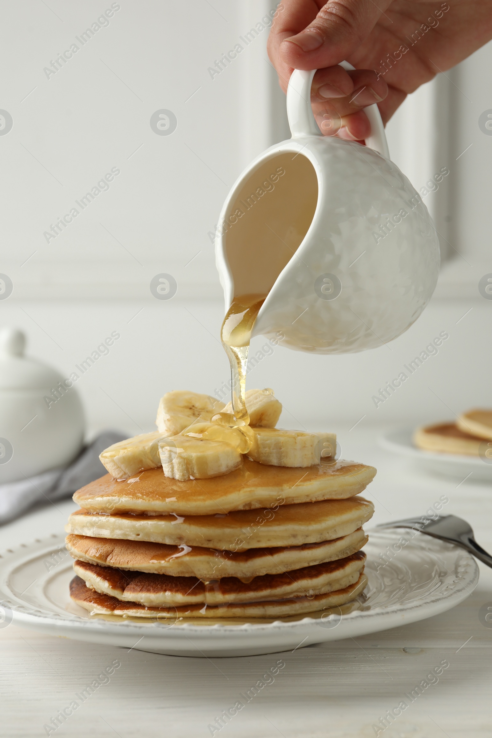 Photo of Woman pouring honey from jug onto delicious pancakes with bananas at white wooden table, closeup