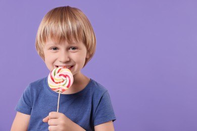 Happy little boy licking colorful lollipop swirl on violet background, space for text