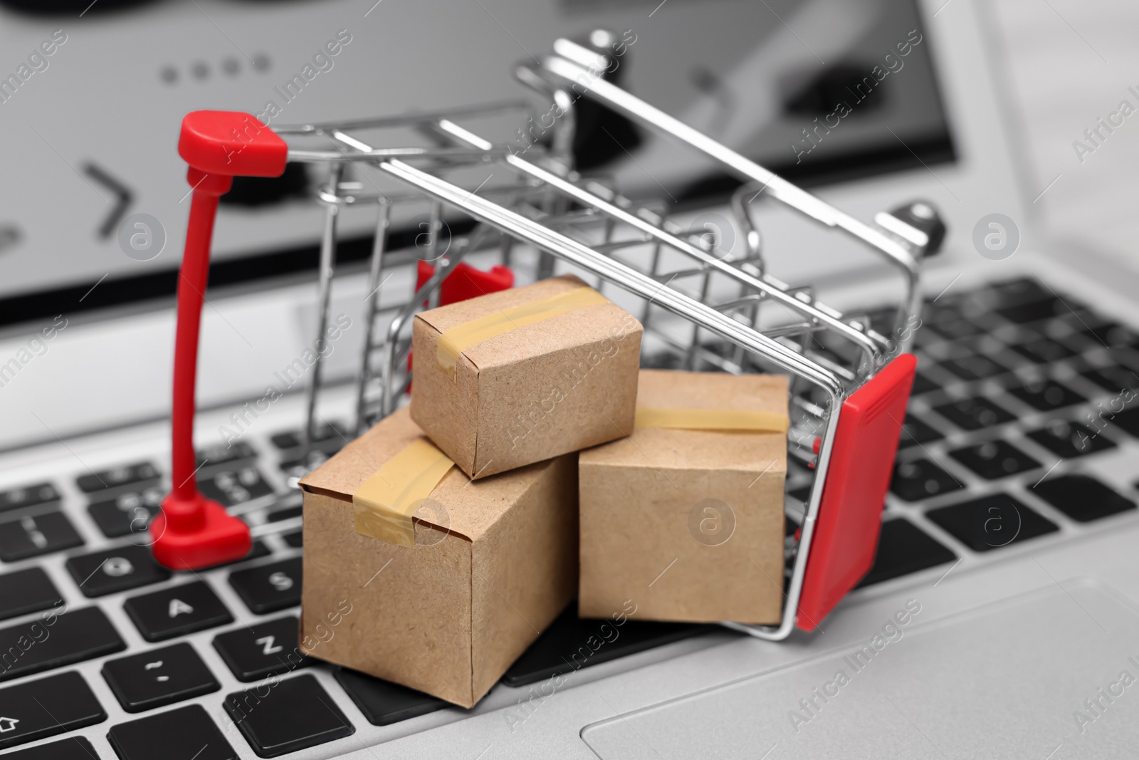 Photo of Internet store. Small cardboard boxes and shopping cart on laptop, closeup