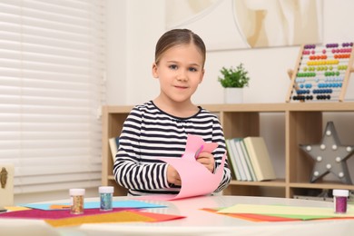 Photo of Cute little girl cutting pink paper at desk in room. Home workplace