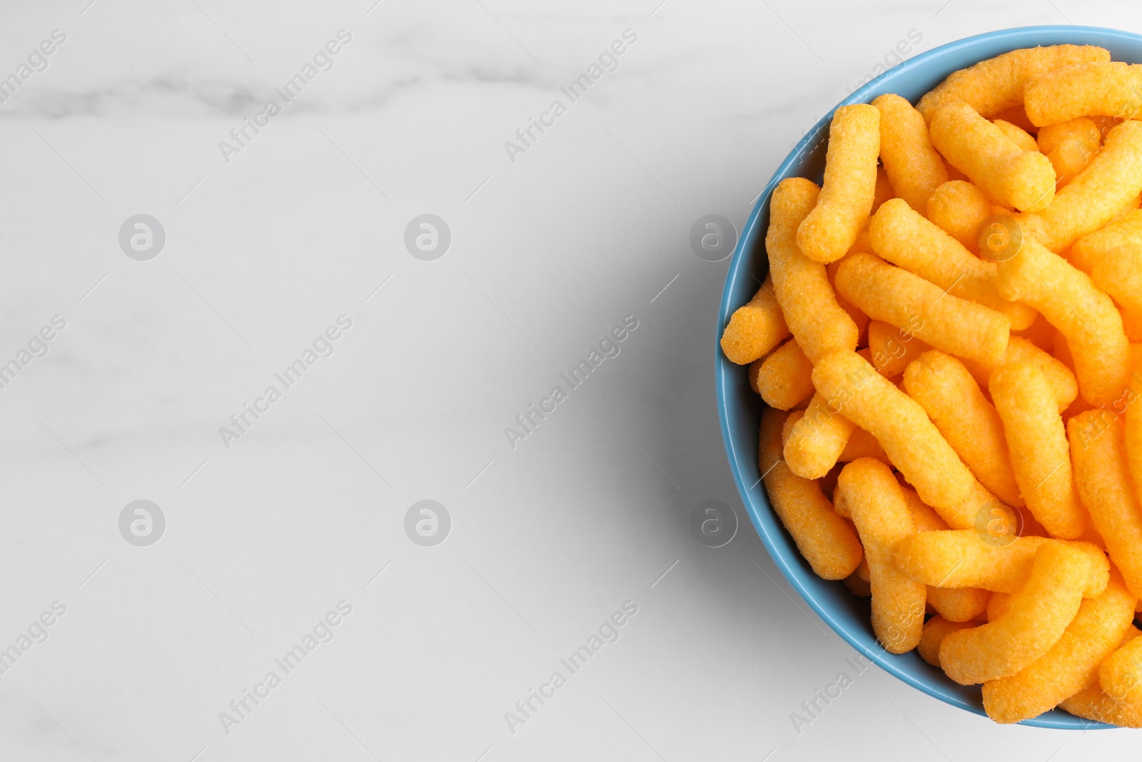 Photo of Bowl of tasty cheesy corn puffs on white marble table, top view. Space for text