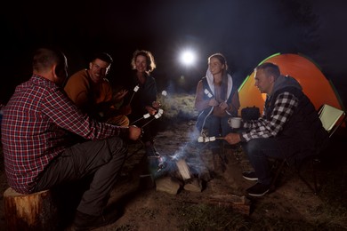 Photo of Group of friends roasting marshmallows on bonfire at camping site in evening