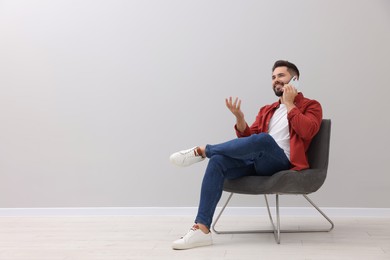 Photo of Handsome man talking on smartphone while sitting in armchair near light grey wall indoors, space for text