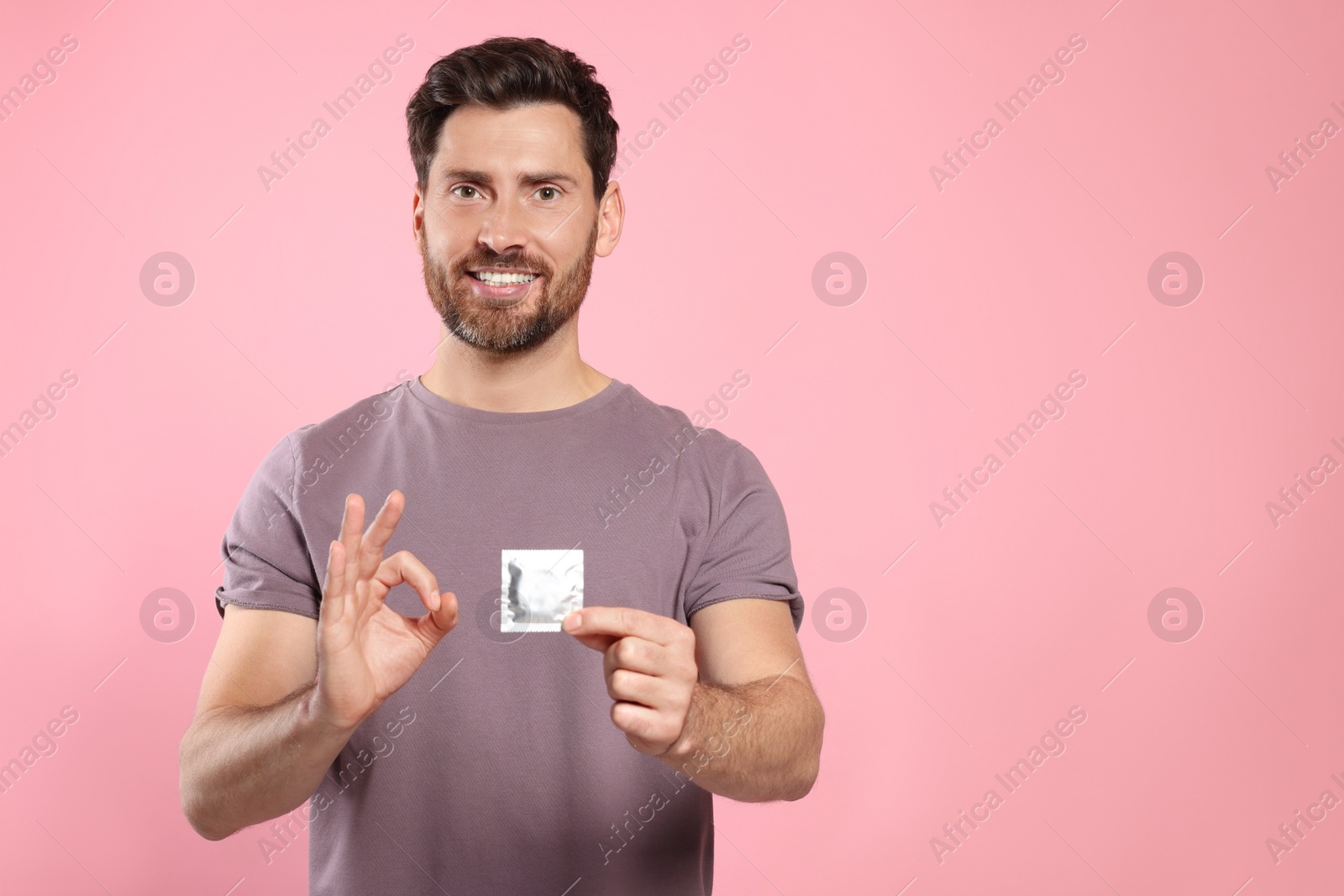 Photo of Man with condom showing ok gesture on pink background, space for text. Safe sex