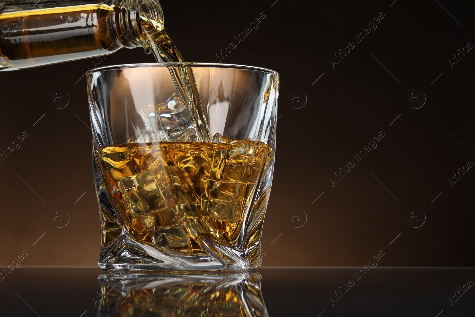 Photo of Pouring whiskey from bottle into glass with ice cubes at table against brown background, space for text