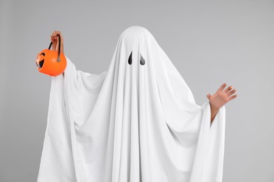 Photo of Woman in white ghost costume holding pumpkin bucket on light grey background. Halloween celebration