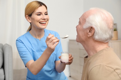 Photo of Nurse feeding elderly man with yogurt indoors. Assisting senior people