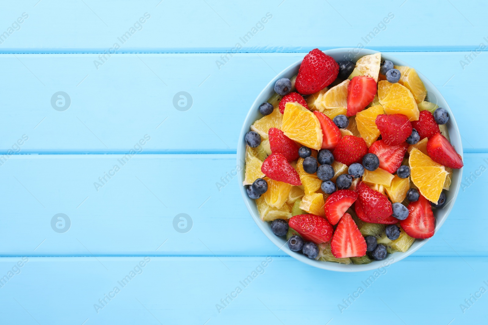 Photo of Delicious fresh fruit salad in bowl on light blue wooden table, top view. Space for text