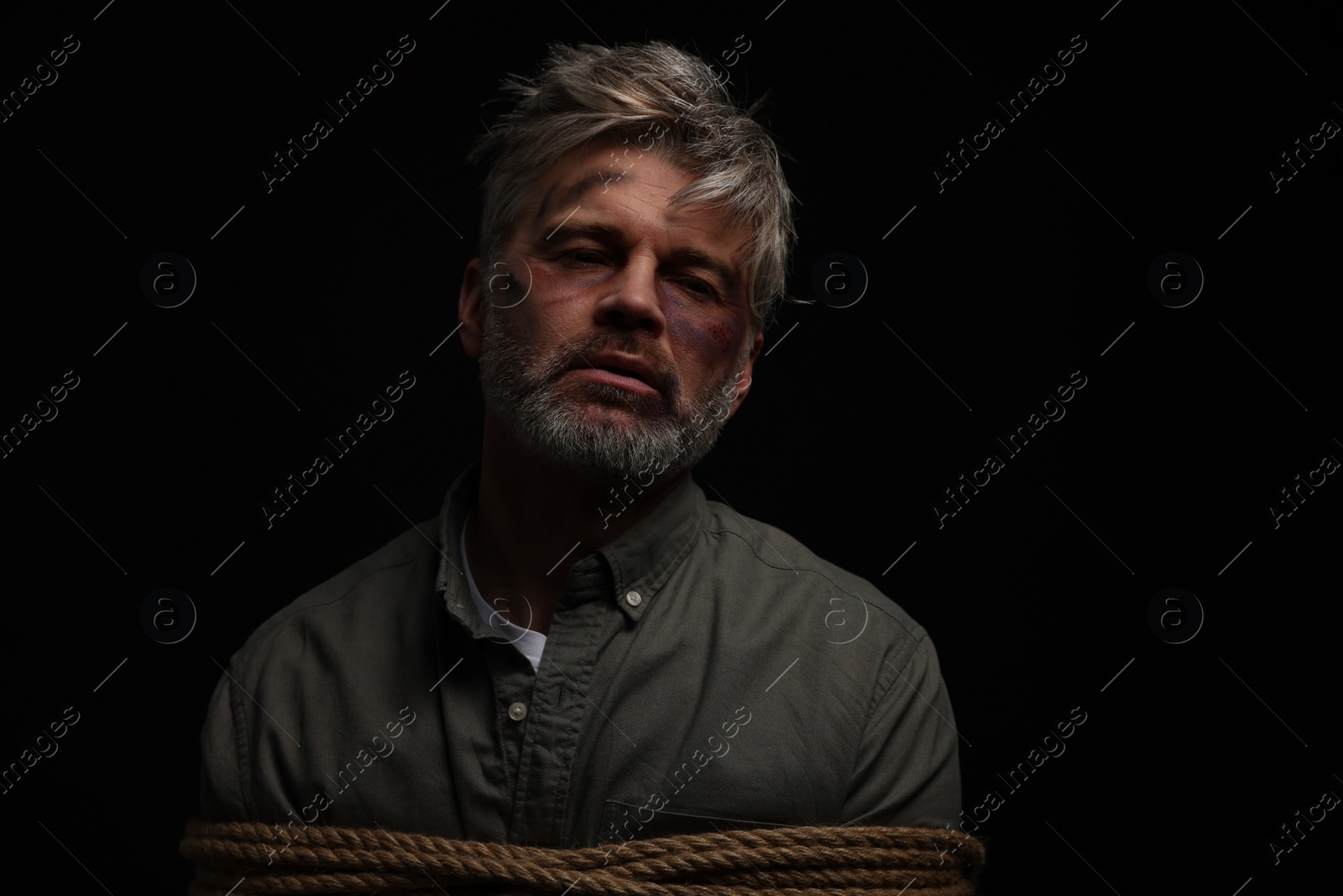 Photo of Beaten man tied with rope on black background. Hostage taking