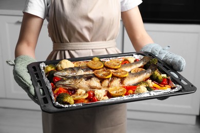 Photo of Woman holding baking tray with sea bass fish and vegetables in kitchen, closeup