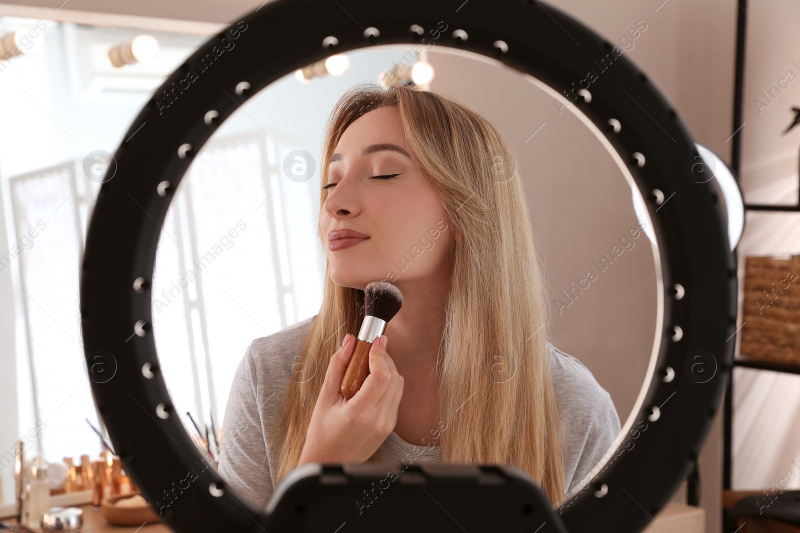 Photo of Beautiful young woman applying face powder with brush indoors, view through ring lamp